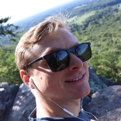 Andrew Akers. Blonde hair, wearing sunglasses, looking back at the camera from on top of a mountain.